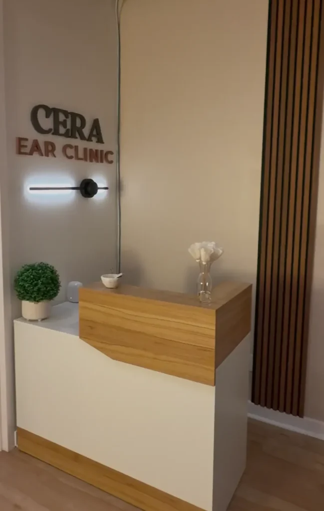 Reception area of CERA Ear Clinic with a small wooden and cream-colored desk, a potted plant, and a glass vase with white flowers, featuring wall signage for the clinic.